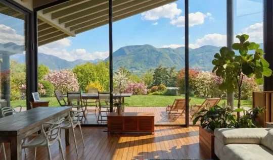 Soggiorno moderno con ampie vetrate vista montagne, patio in legno e giardino primaverile con alberi in fiore.