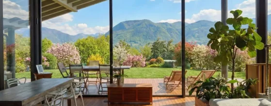 Soggiorno moderno con ampie vetrate vista montagne, patio in legno e giardino primaverile con alberi in fiore.