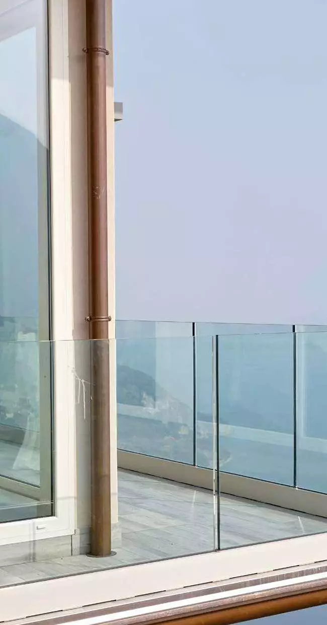 Dettaglio di un angolo di balcone con parapetto in vetro trasparente, vetrata a tutta altezza e pluviale in rame.