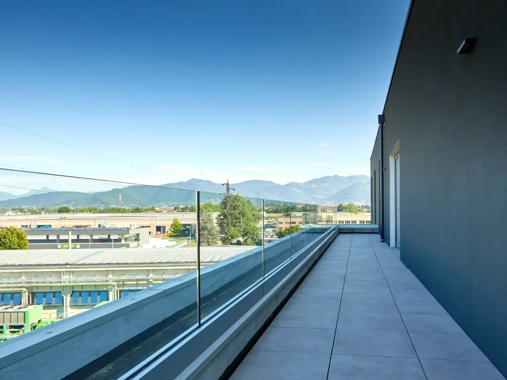 Parapetto in vetro Garda ONE con fissaggio sopra pavimento su lunga terrazza esterna con piastrelle grigie.