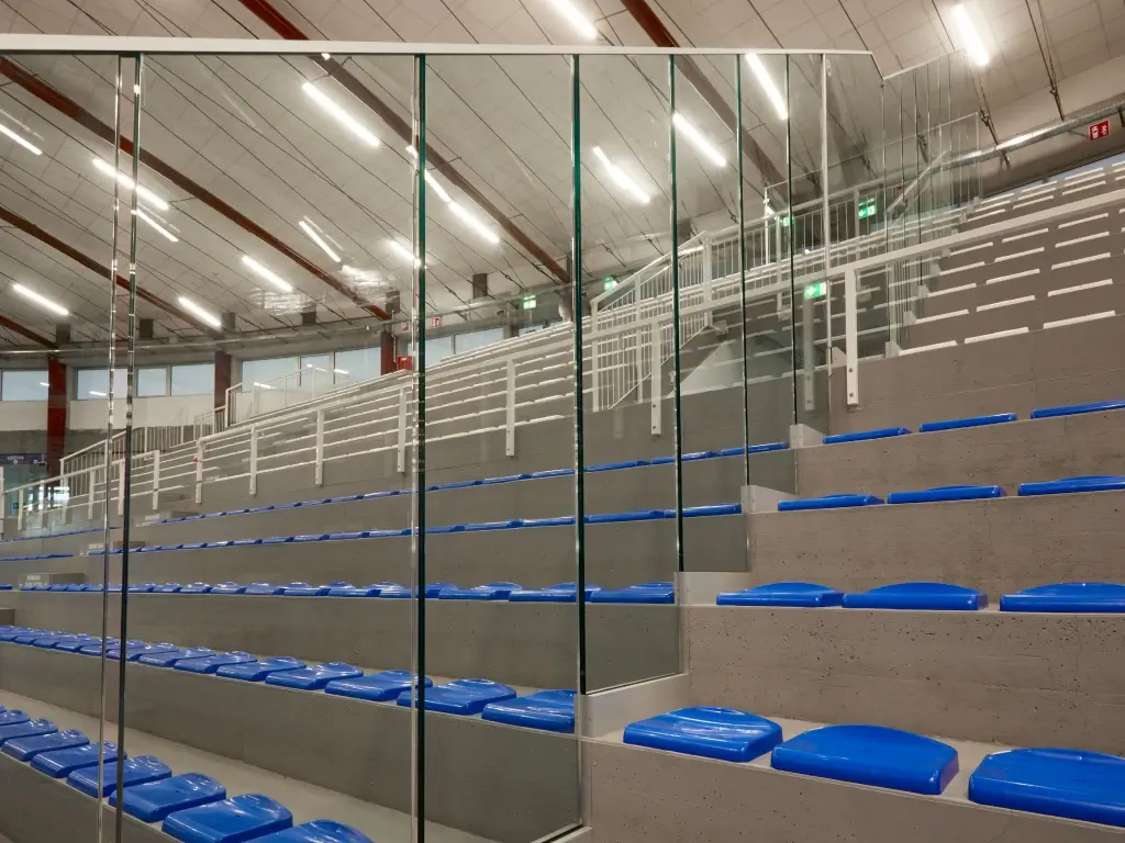 Barriera di sicurezza in vetro Garda ONE su gradinate di un impianto sportivo indoor con sedute blu.